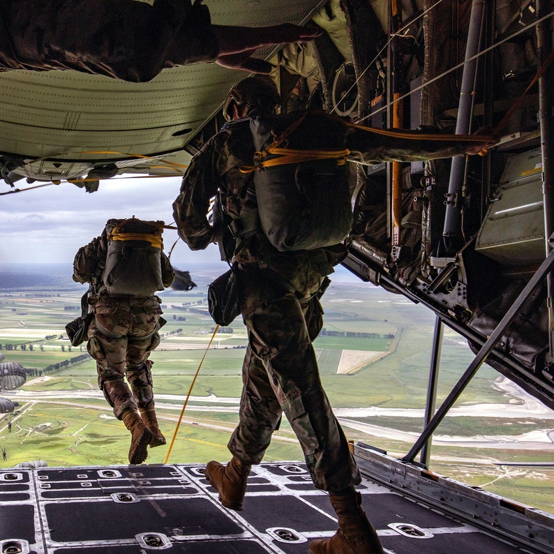 May include: Two people in military uniforms are preparing to jump out of an airplane. They are wearing parachutes and are looking out of the open cargo door. The sky is cloudy and the ground is visible below.