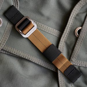 May include: Close-up of a black and tan strap with a metal buckle. The strap is made of a woven material and features the text "HAWKRIGGER" on the buckle. The background is a textured, olive green fabric.
