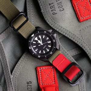 Peut inclure: Une montre analogique noire avec un cadran rond, des index blancs et le mot QUARTZ imprimé sur le cadran. La montre présente une lunette rotative et un bracelet en nylon passant du vert olive à une section rouge vif avec des boucles noires.