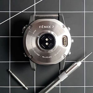 Puede incluir: Un reloj inteligente plateado con una esfera negra y el texto "FENIX 7" en el bisel. El reloj está sobre una superficie negra con una pequeña herramienta plateada.