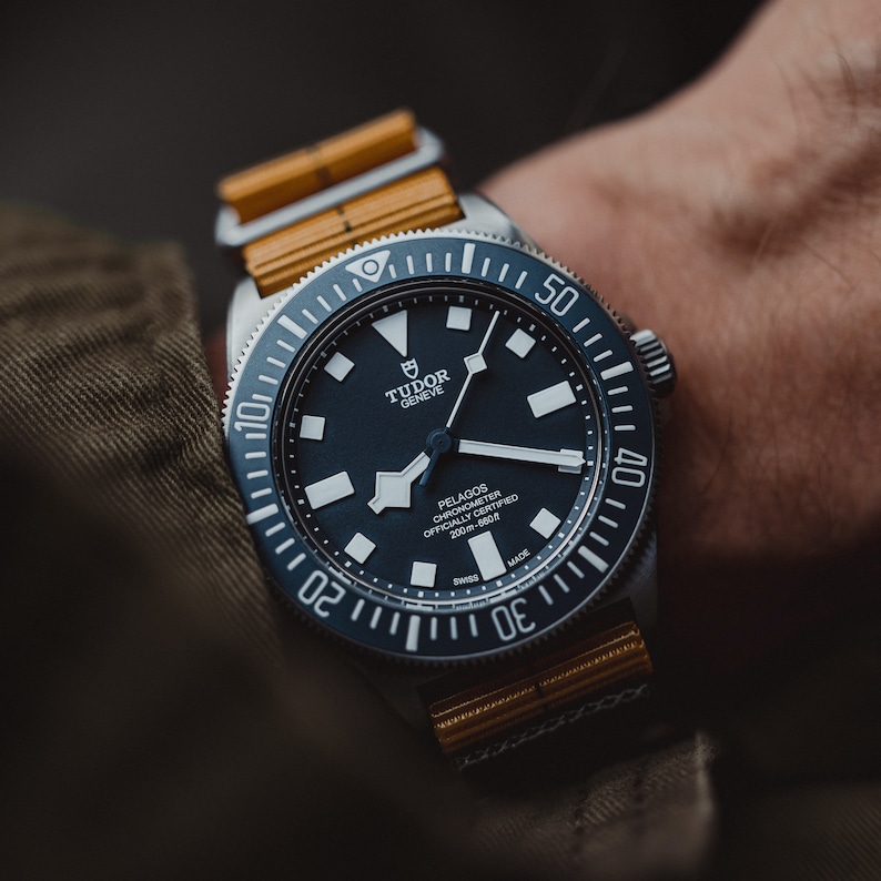 May include: A black Tudor Pelagos Chronometer watch with a black face and white markings. The watch has a yellow and brown fabric strap and a rotating bezel with black markings.