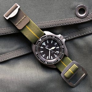 May include: A wristwatch with a black face and a green and yellow nylon strap. The watch has a silver-colored bezel with white markings and a silver-colored case. The strap is secured with a silver-colored buckle.