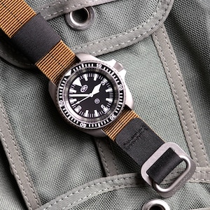 May include: A stainless steel wristwatch with a black face and a tan and black nylon strap. The watch has a rotating bezel and is set against a green fabric background.