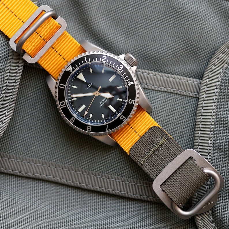 May include: A black and silver wristwatch with a yellow and green nylon strap. The watch face has a black background with white markings and the word "MILK" printed on it. The watch has a date window at the 3 o'clock position.