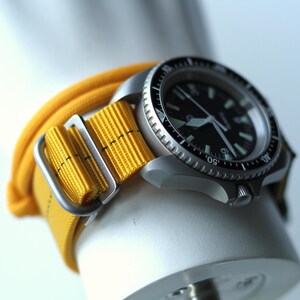 May include: A silver wristwatch with a black face and a yellow nylon strap. The watch has a rotating bezel with numbers and markings.