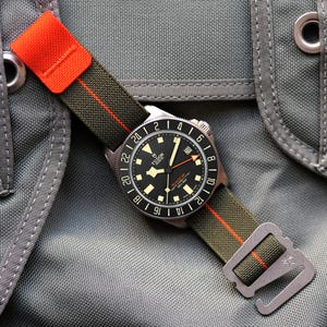 Puede incluir: Reloj de pulsera con esfera negra y correa de tela verde con una raya naranja. El reloj tiene números y agujas blancas, y la marca "TUDOR" es visible. La correa se sujeta con un cierre metálico.