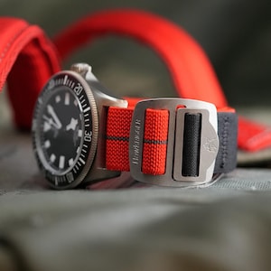 May include: Close-up of a wristwatch with a black face and a silver bezel. The watch has a red and black nylon strap with a silver buckle. The strap is marked with the text "HAWKRIGGER".