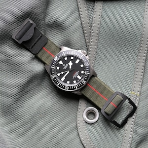 Op de afbeelding: Een horloge met een zwarte wijzerplaat en een groene band met een rode streep. Het horloge heeft een zilveren lunette en een zwarte gesp. Het horloge wordt gepresenteerd op een gestructureerde, grijze stof.