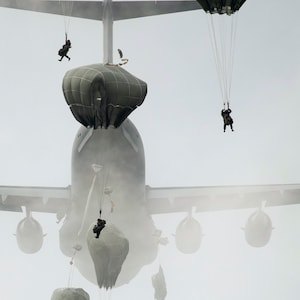 May include: A military cargo plane releases parachutes with soldiers descending from the sky. The plane is partially obscured by fog or clouds.