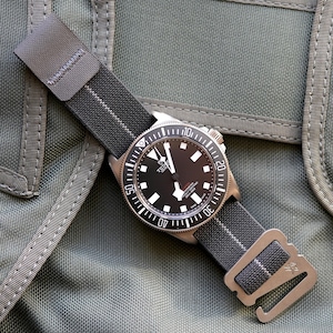 Könnte beinhalten: Eine Armbanduhr mit schwarzem Zifferblatt und grau-weiß gestreiftem Armband. Die Uhr hat eine silberne Lünette mit weißen Markierungen und ein silbernes Gehäuse. Das Armband wird mit einer silbernen Schließe befestigt. Grüner, strukturierter Hintergrund.