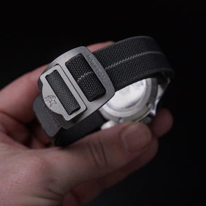 May include: A close-up shot of a grey watch strap with a metal clasp. The strap is made of a textured nylon material with a white stripe. The clasp is engraved with the text "HawkRigger" and a star logo. The watch face is visible.