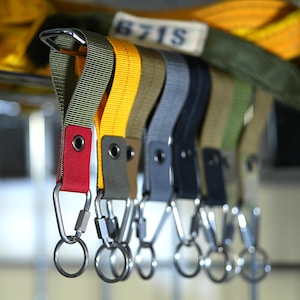 Pode incluir: Um conjunto de sete chaveiros coloridos com argolas e clipes de metal prateado. Os chaveiros são feitos de tecido tecido em amarelo, verde, azul, cinza e preto. Os chaveiros são etiquetados com o número "6715".