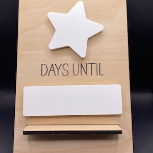 May include: A wooden countdown board with a white star and a blank space for writing in the number of days until a special event. The text "DAYS UNTIL" is written on the board.