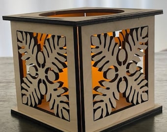 May include: A wooden candle holder with a square shape and intricate cut-out designs of tropical leaves. The candle holder is lit with a warm orange glow.