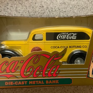 May include: A yellow Coca-Cola die-cast metal bank in its original box. The bank is a vintage-style delivery truck with black wheels and the Coca-Cola logo. The box features the Coca-Cola logo and an image of a woman drinking Coca-Cola.