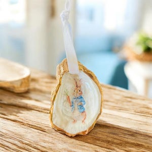 May include: An oyster shell ornament featuring a watercolor illustration of Peter Rabbit holding a carrot. The shell has a gold-colored edge and a white ribbon for hanging. The ornament is on a wooden surface.