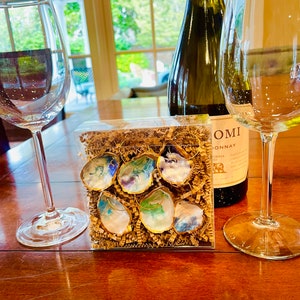 May include: A set of six painted oyster shell earrings in a clear plastic box with brown packing paper. The earrings are painted with various shades of blue, green, and purple. The box is sitting on a wooden table next to a bottle of white wine and two wine glasses.
