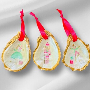 May include: Three gold-painted oyster shell ornaments with pink ribbon hangers. Each shell features a hand-painted illustration of a different character from the Nutcracker ballet: a ballerina, a nutcracker soldier, and a mouse king.