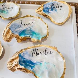 May include: Four gold-painted oyster shells with blue and white marbled interiors. Each shell has a name written in black calligraphy: Sunny, Betsy, Jeannie, and Marilee.
