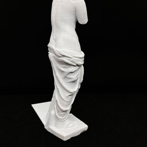 May include: White 3D printed statue of the Venus de Milo, a famous Greek sculpture. The statue is posed with its head tilted down and its arms missing. The statue is standing on a white base.