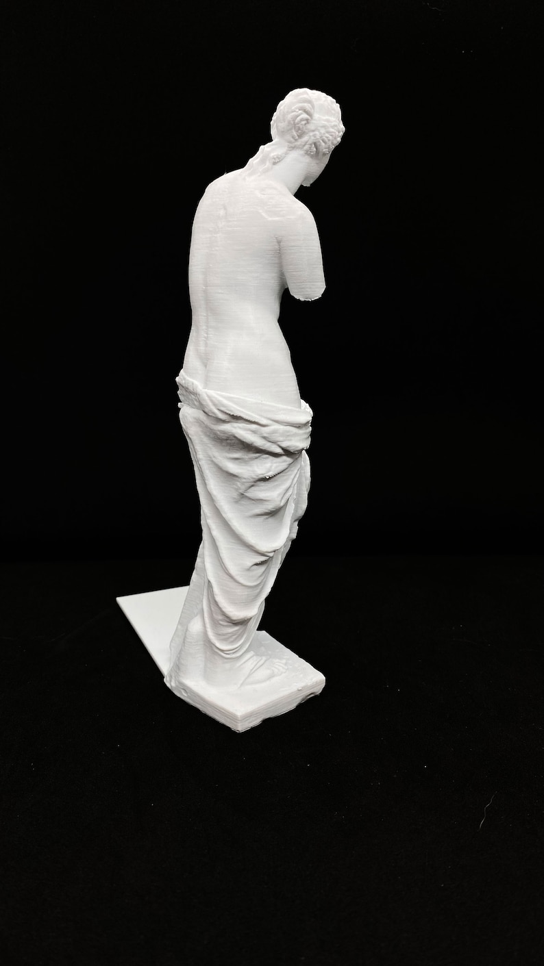 May include: White 3D printed statue of the Venus de Milo, a famous Greek sculpture. The statue is posed with its head tilted down and its arms missing. The statue is standing on a white base.