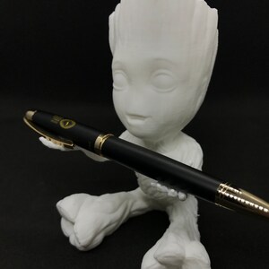 Peut inclure: Figurines Groot en 3D imprimé en blanc tenant un stylo noir avec des accents dorés. Le stylo porte l'inscription "The Pen Company".