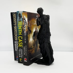 May include: A black bookend in the shape of the Venus de Milo statue holding up a stack of four books. The books are titled "The Tenth Case", "As Esu Legenda", "As Esu Legenda", and "Sidney Sheldon - Memories of Midnight".