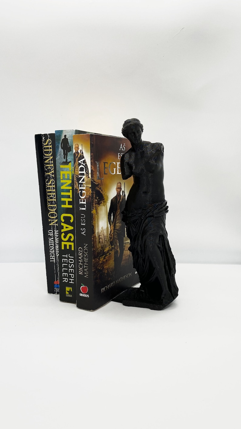 May include: A black bookend in the shape of the Venus de Milo statue holding up a stack of four books. The books are titled "The Tenth Case", "As Esu Legenda", "As Esu Legenda", and "Sidney Sheldon - Memories of Midnight".