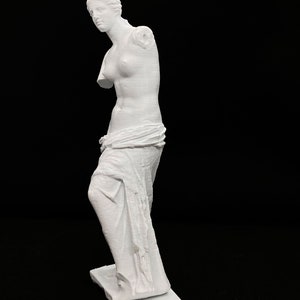 May include: A white 3D printed statue of the Venus de Milo, a famous Greek sculpture. The statue is posed with one arm missing and a draped cloth around the waist.