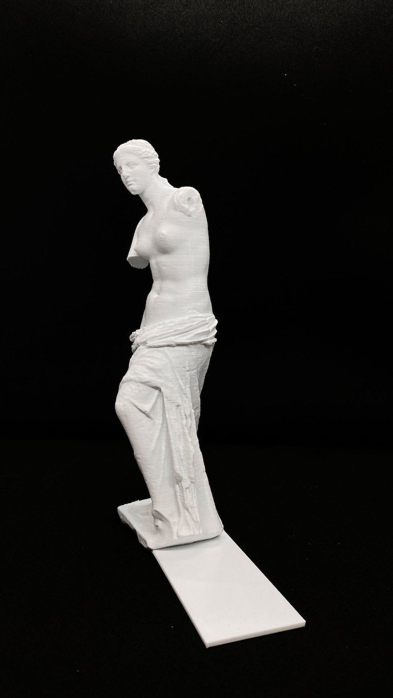 May include: A white 3D printed statue of the Venus de Milo, a famous Greek sculpture. The statue is posed with one arm missing and a draped cloth around the waist.