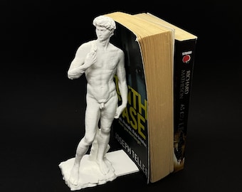 May include: White plastic bookend featuring a statue of a nude male figure. The bookend is holding up a book titled "The Fifth Witness" by Joseph Teller.