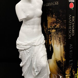 May include: A white 3D printed statue of the Venus de Milo, a famous Greek sculpture, is used as a bookend. The statue is holding a book titled "The Legend As Easy As E.B.U." by Richard Matheson.