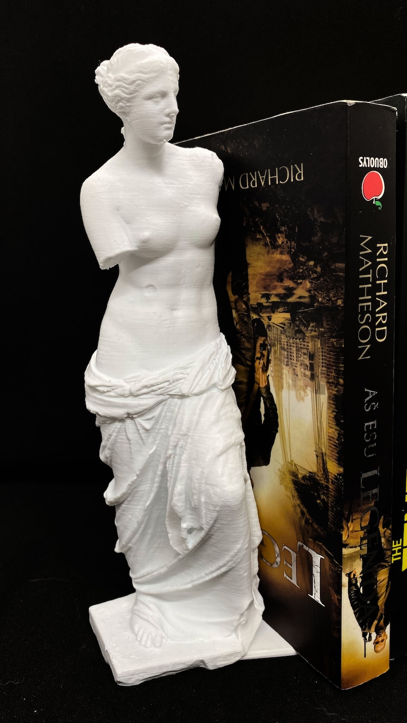 May include: A white 3D printed statue of the Venus de Milo, a famous Greek sculpture, is used as a bookend. The statue is holding a book titled "The Legend As Easy As E.B.U." by Richard Matheson.