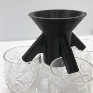 Op de afbeelding: Een zwart 3D-geprint apparaat met een trechtervormige bovenkant en drie poten. Het apparaat is geplaatst boven vier heldere glazen bekers met een decoratief patroon.