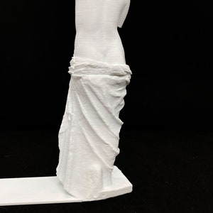 May include: White 3D printed statue of a woman standing with her arms crossed and a draped cloth around her waist.
