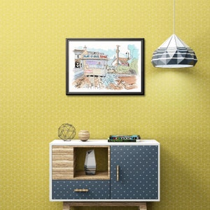 May include: A framed watercolour painting of a seaside town, featuring a shop sign that reads "Lots of Great Fudge". The artwork is displayed above a modern cabinet with geometric patterns, a decorative lamp, and a yellow patterned wall.