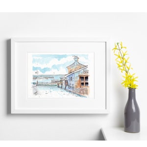 May include: Watercolour painting of a seaside shop with the word "BIK" visible. The shop is painted in shades of blue and brown, with a bicycle parked outside. A white frame surrounds the artwork. A vase with yellow flowers is on the right.