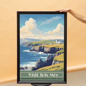 May include: A framed travel poster featuring a coastal scene with cliffs, the sea, and a bright sky with clouds. The poster has the text "Visit Inis Mór" at the bottom, inviting viewers to explore the location.