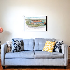May include: A framed watercolour painting depicting a rural building with a red roof and stone walls. The artwork is displayed above a grey sofa with patterned throw cushions in blue and yellow.