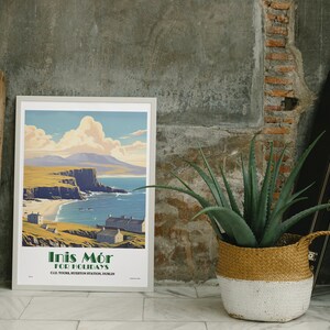 May include: A framed travel poster for Inis Mór for Holidays, depicting a coastal scene with a beach, cliffs, and buildings. The poster has a white border and is propped against a grey wall. A wooden crate and a potted aloe vera plant are also in the image.