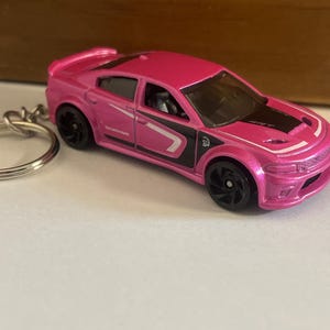 May include: A pink Dodge Charger toy car keychain. The car has black wheels and a black stripe on the hood. The keychain ring is silver.