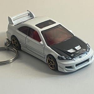 May include: A white toy car with a black hood and a rear spoiler. The car has gold-colored wheels and a red interior. Attached to the car is a silver key ring.