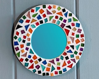 May include: A round mirror with a white frame decorated with colorful mosaic tiles. The mirror has a turquoise blue reflective surface.