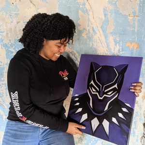 May include: A person is holding a purple canvas painting of Black Panther. The painting is a close-up of Black Panther's face, with a black mask and white accents. The person is wearing a black hoodie with the word "Pink" on the front and "PINK" on the sleeve.