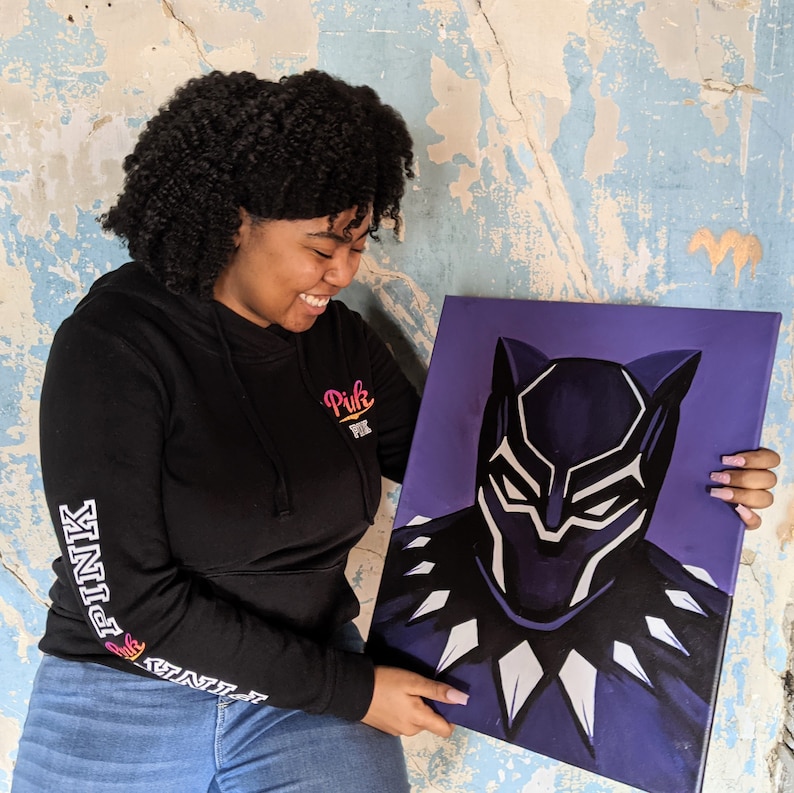 May include: A person is holding a purple canvas painting of Black Panther. The painting is a close-up of Black Panther's face, with a black mask and white accents. The person is wearing a black hoodie with the word "Pink" on the front and "PINK" on the sleeve.