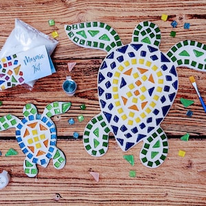 May include: A mosaic kit with two turtle shapes, one large and one small. The turtles are made of white ceramic and decorated with blue, green, yellow, and orange mosaic tiles. The kit includes a bag of white glue, a small bottle of blue glue, a paintbrush, and extra mosaic tiles.