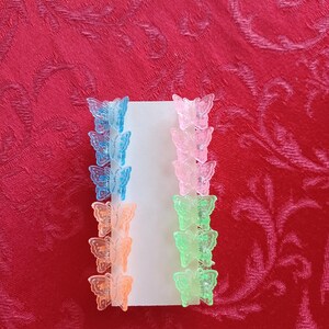 May include: A collection of small butterfly hair clips in various colors, including blue, pink, orange, and green. The clips are arranged on a white card against a red background. These accessories are ideal for adding a touch of whimsy to any hairstyle.