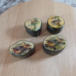 May include: Four small decorative boxes with bird and floral designs. One is heart-shaped, the others are oval or round. The boxes have a dark, textured exterior and a yellow or cream-colored lid with a detailed illustration of birds or flowers.