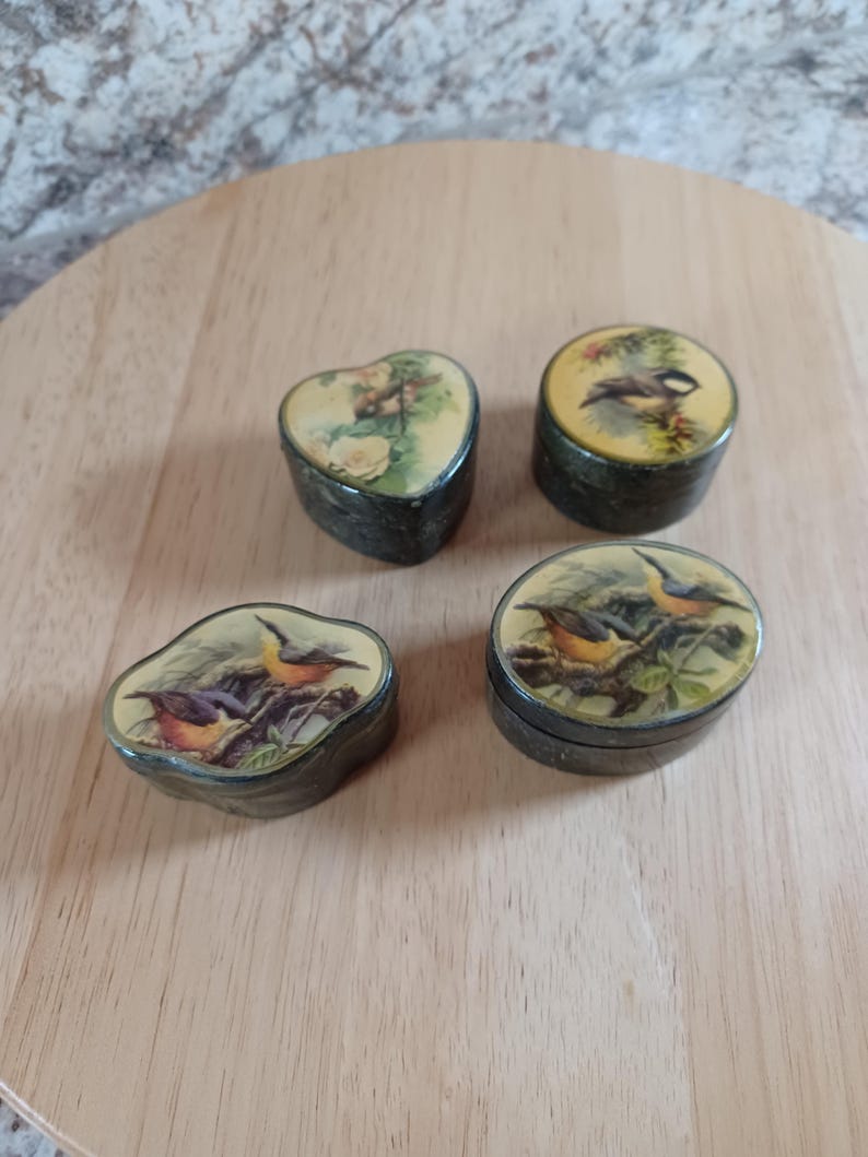 May include: Four small decorative boxes with bird and floral designs. One is heart-shaped, the others are oval or round. The boxes have a dark, textured exterior and a yellow or cream-colored lid with a detailed illustration of birds or flowers.