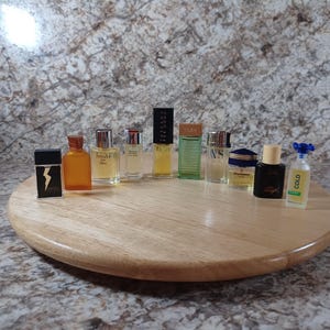 May include: A collection of miniature perfume bottles of various shapes and colors, arranged on a round wooden tray. The bottles include amber, clear, and black containers, some with gold accents. The background is a speckled, neutral-toned surface.